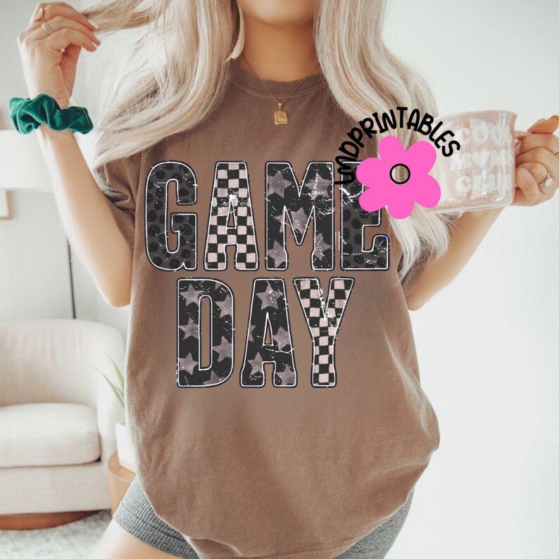 May include: A brown t-shirt with the words "GAME DAY" in a distressed, layered design. The letters feature a mix of patterns, including checkered, leopard print, and stars. The shirt has a casual, relaxed fit.