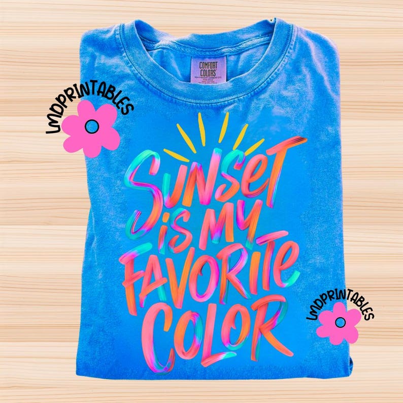 May include: A blue Comfort Colors t-shirt with the text "Sunset is my favorite color" in colorful, layered lettering. The shirt has a crew neck and is folded. The design includes a sun graphic.