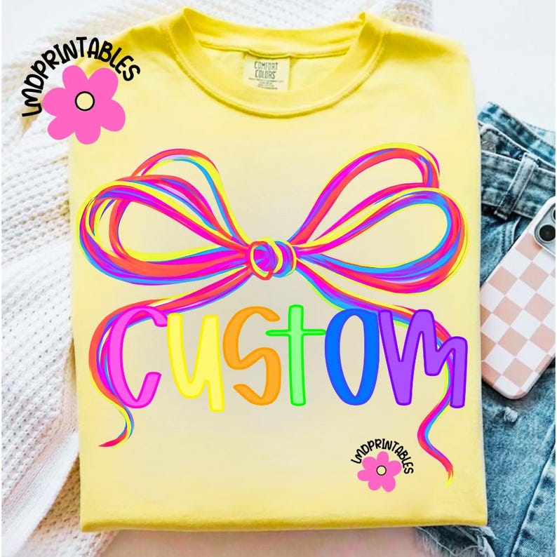 May include: Yellow t-shirt with a colorful bow design and the word "CUSTOM" in rainbow colors. The shirt has a crew neck and short sleeves. The text "LMDPRINTABLES" is printed on the shirt.