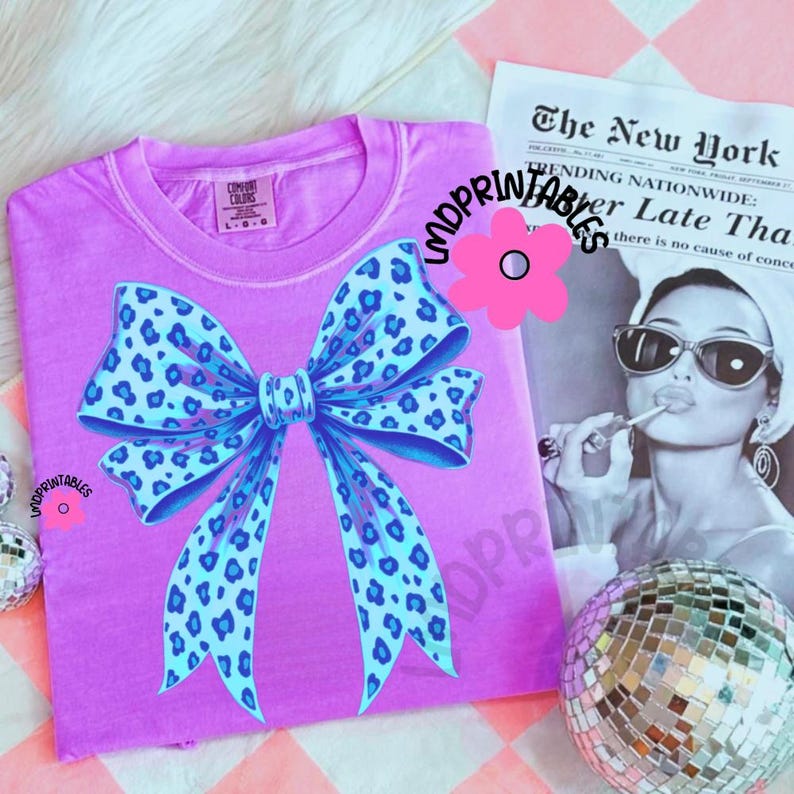May include: A lilac-colored t-shirt featuring a large bow design with a blue leopard print. The shirt has a crew neck and short sleeves. The text "LMDPRINTABLES" is visible. A newspaper and disco ball are in the background.