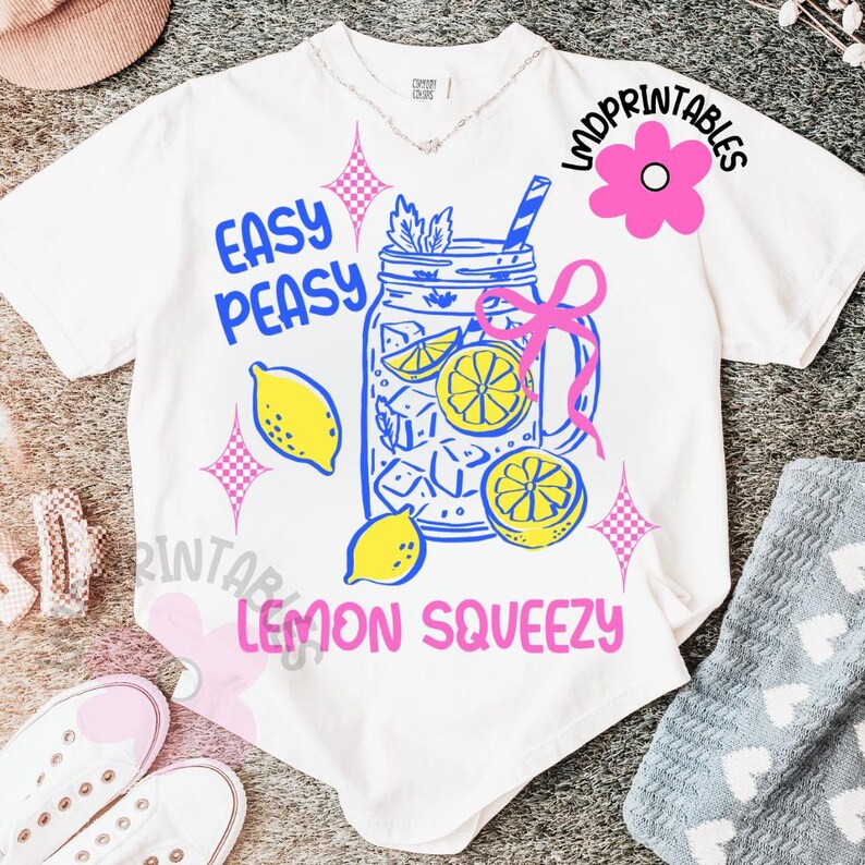 May include: White t-shirt featuring a blue and pink graphic of a lemon drink in a mason jar. The text on the shirt reads "Easy Peasy Lemon Squeezy" with a pink flower and starbursts. The shirt is on a light gray surface.