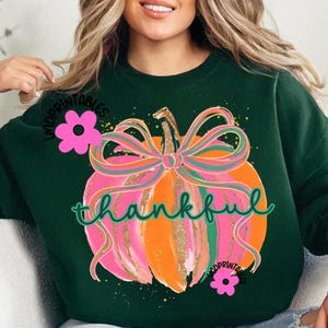 Peut inclure: Sweat-shirt vert fonc&eacute; avec un motif de citrouille color&eacute;e. La citrouille est orange et rose avec une tige et un n&oelig;ud dor&eacute;s &agrave; paillettes. Le mot "thankful" est &eacute;crit en vert. Des fleurs roses et le texte "MDPRINTABLES" sont &eacute;galement visibles.