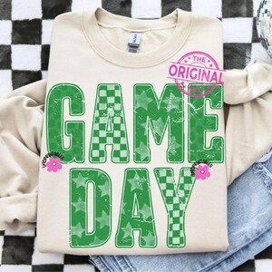 May include: Cream-colored sweatshirt with the words "GAME DAY" in distressed green lettering. The letters feature a mix of star and checkered patterns. A pink floral graphic and the words "THE ORIGINAL" are also visible.