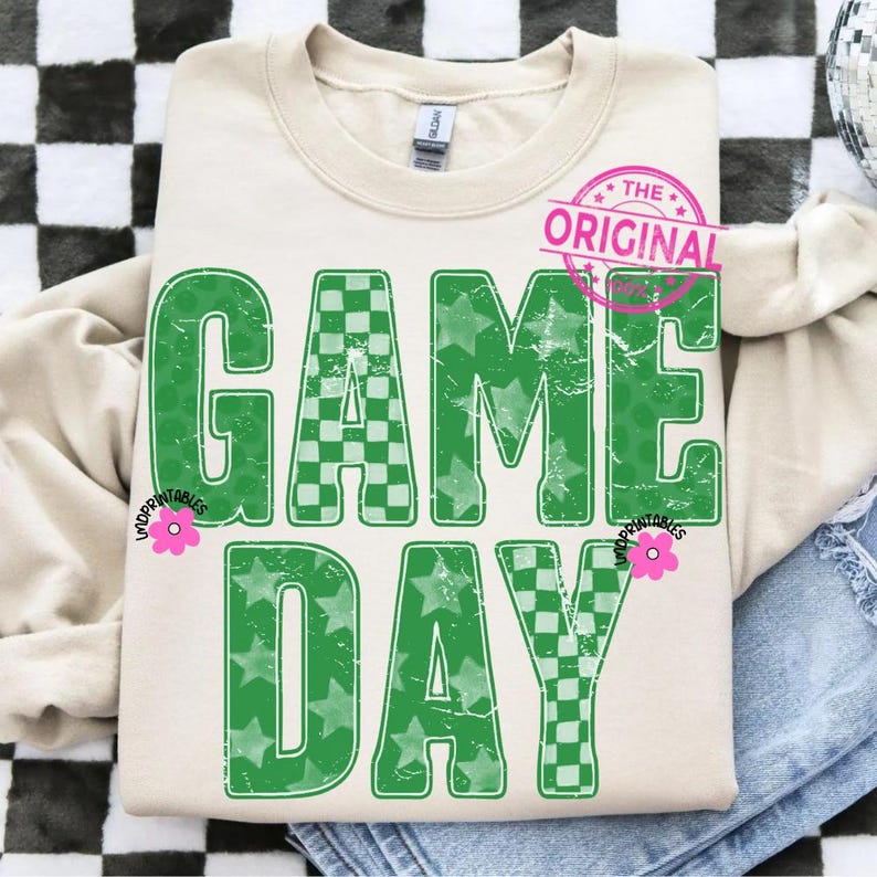 May include: Cream-colored sweatshirt with the words "GAME DAY" in distressed green lettering. The letters feature a mix of star and checkered patterns. A pink floral graphic and the words "THE ORIGINAL" are also visible.