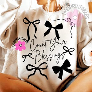 K&ouml;nnte beinhalten: Cremefarbenes Sweatshirt mit schwarzen Schleifen-Grafiken und dem Schriftzug "Count Your Blessings" in einem stilvollen Schriftzug. Das Design umfasst mehrere Schleifen-Illustrationen und schafft ein optisch ansprechendes und inspirierendes Kleidungsst&uuml;ck.