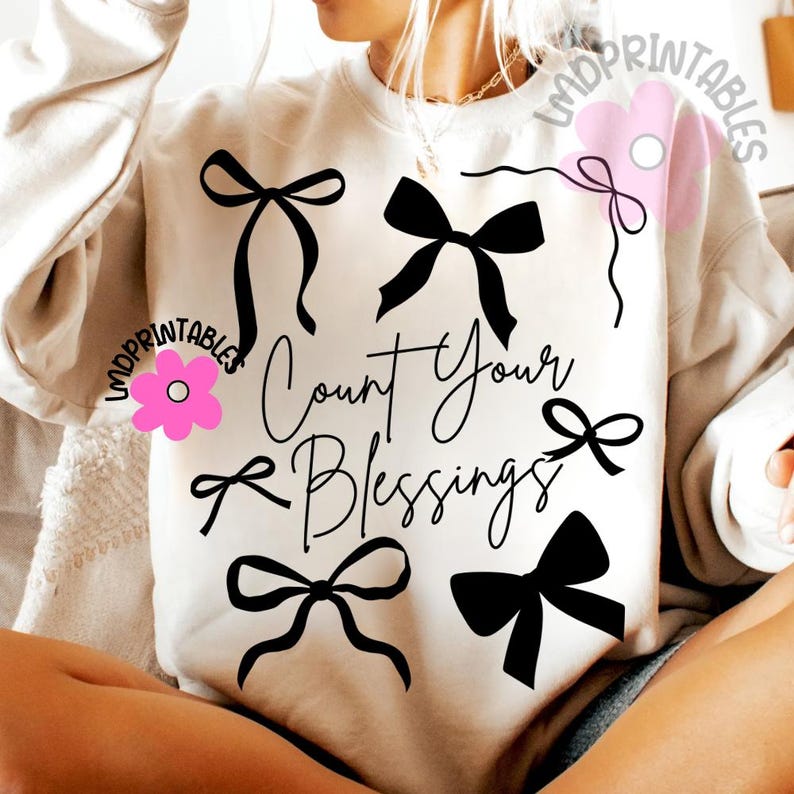 K&ouml;nnte beinhalten: Cremefarbenes Sweatshirt mit schwarzen Schleifen-Grafiken und dem Schriftzug "Count Your Blessings" in einem stilvollen Schriftzug. Das Design umfasst mehrere Schleifen-Illustrationen und schafft ein optisch ansprechendes und inspirierendes Kleidungsst&uuml;ck.