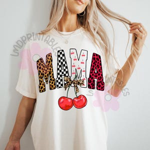 May include: Cream-colored t-shirt with the word "MAMA" in a bold, playful design. The letters feature leopard print, a checkered pattern, cherries, and a pink leopard print. A bow and two red cherries complete the design.