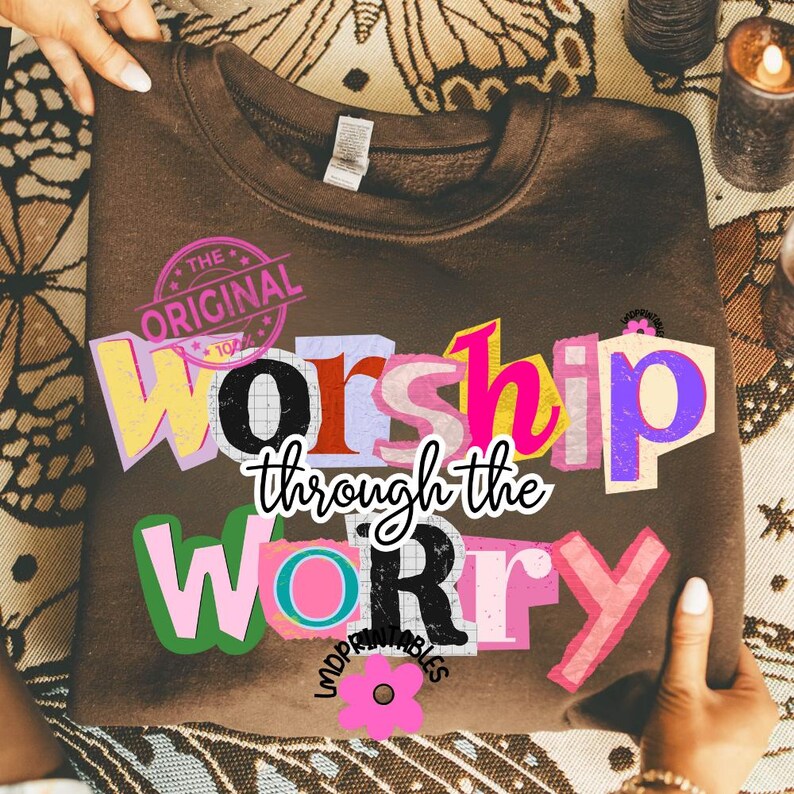 May include: A brown sweatshirt with the words "Worship through the Worry" in a colorful, collage-style design. The text includes the words "The Original" in a circular stamp. A pink flower graphic is also visible.