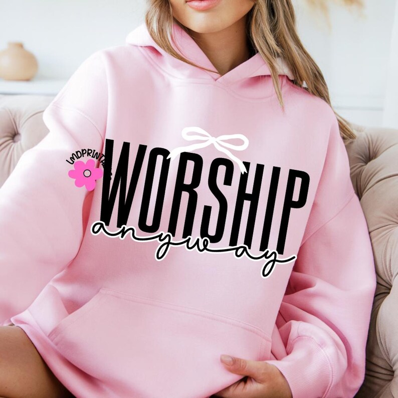 May include: A pale pink hoodie featuring the text "WORSHIP anyway" in black and white. The hoodie has a front pocket and a white bow graphic. A pink flower logo is on the left sleeve. The hoodie is a casual garment.