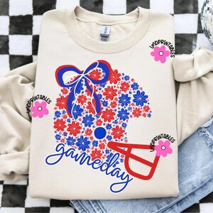 May include: Cream-colored sweatshirt featuring a football helmet design made of red and blue flowers. The word "gameday" is written in cursive below the helmet. The design includes a bow and a circular detail.