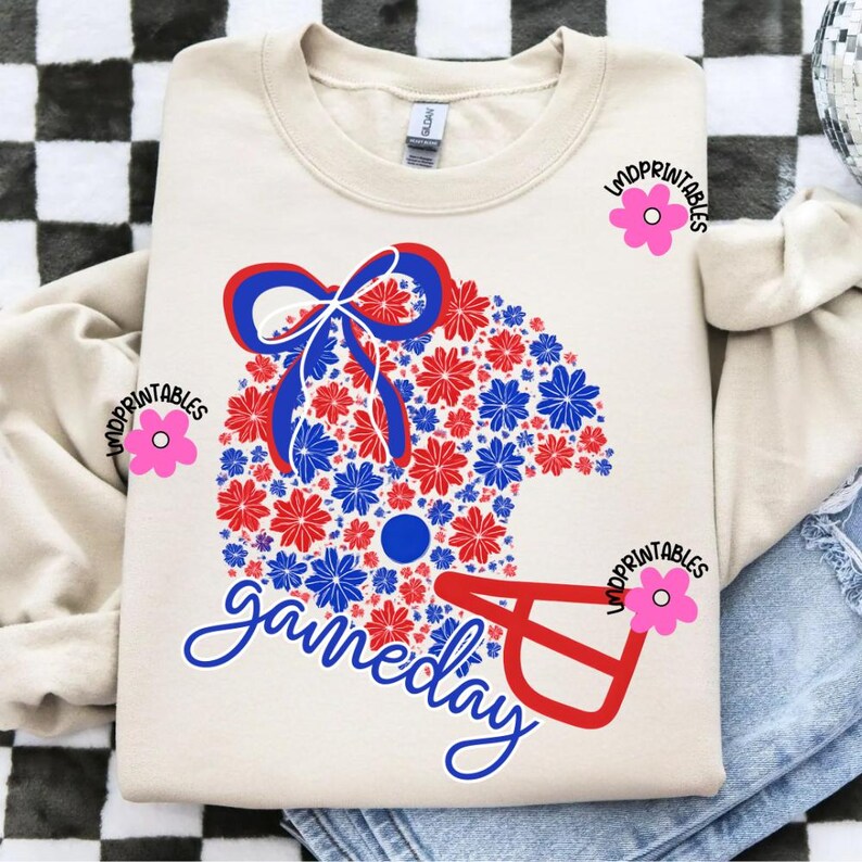 May include: Cream-colored sweatshirt featuring a football helmet design made of red and blue flowers. The word "gameday" is written in cursive below the helmet. The design includes a bow and a circular detail.