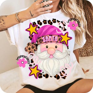 Op de afbeelding: Wit T-shirt met een kerstman-afbeelding. Het ontwerp heeft een roze hoed met het woord "SANTA", een witte baard en een roze neus. Het ontwerp is omgeven door gele sterren, roze bloemen en luipaardprint accenten.