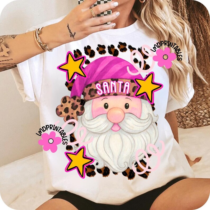 Op de afbeelding: Wit T-shirt met een kerstman-afbeelding. Het ontwerp heeft een roze hoed met het woord "SANTA", een witte baard en een roze neus. Het ontwerp is omgeven door gele sterren, roze bloemen en luipaardprint accenten.