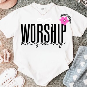 May include: White t-shirt with the words "WORSHIP anyway" in black script. A pink flower graphic is on the upper right, and a white bow is above the word "WORSHIP". The shirt has a V-neck and short sleeves.