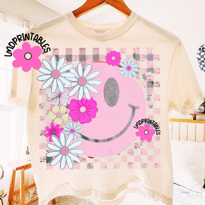 Op de afbeelding: Cr&egrave;mekleurig T-shirt met een grote roze smiley met een madeliefjesontwerp en een geblokte achtergrond. Het shirt heeft de tekst "LMDPrintables" erop. Het ontwerp bevat roze, wit en lichtblauwe kleuren.