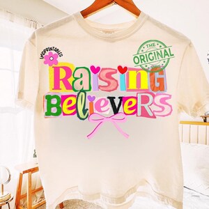May include: Cream-colored t-shirt with the text "Raising Believers" in colorful, layered letters. A pink bow and a flower graphic are also present. The words "The Original" are stamped in green.