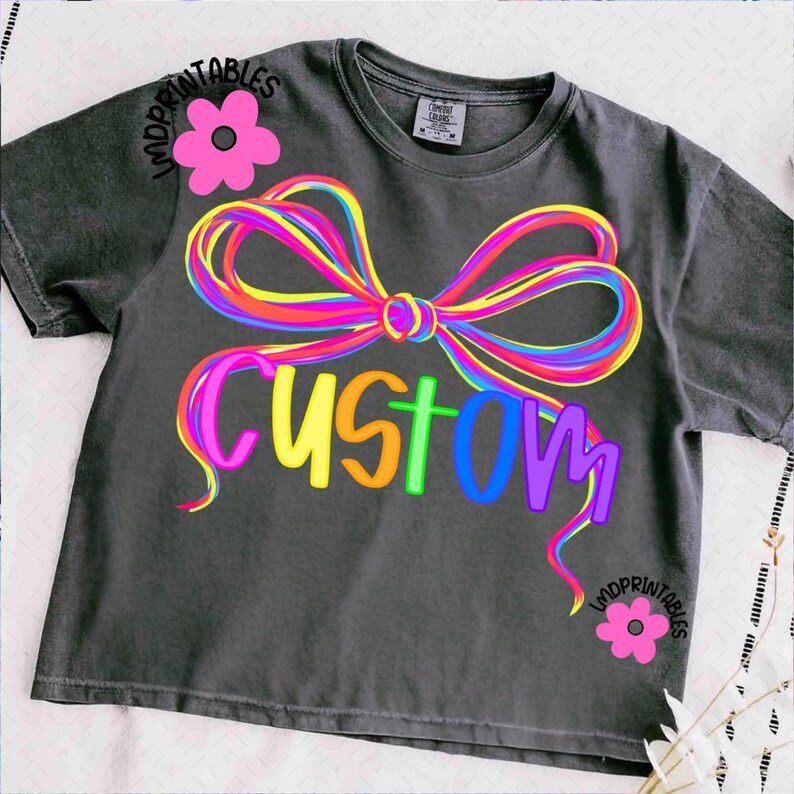 May include: Dark gray t-shirt with a colorful bow design and the word "CUSTOM" in rainbow colors. The shirt also features pink flower graphics and the text "IMDPRINTABLES".