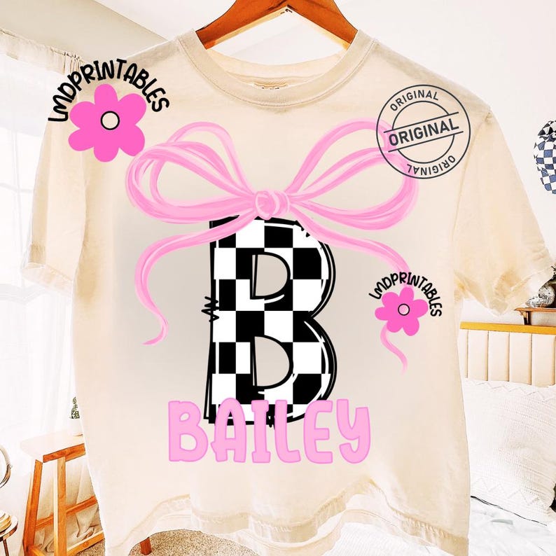 May include: Cream-colored t-shirt featuring a large, black and white checkered letter "B" with a pink bow and the name "Bailey" in pink. The shirt also has pink flower graphics and the word "Original" in a circle.