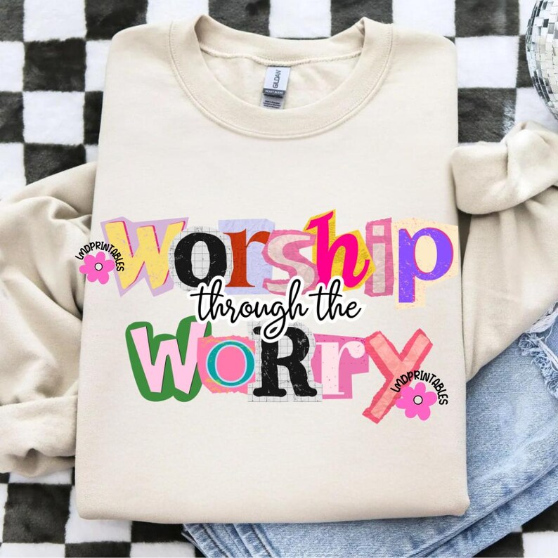 May include: Cream-colored sweatshirt with the text "Worship through the Worry" in a colorful, cut-out style. The design features various colors like pink, yellow, purple, and green. The sweatshirt is folded on a denim surface.