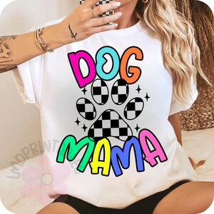 May include: White t-shirt with the words "DOG MAMA" in colorful bubble letters. The design includes a paw print with a checkered pattern and star accents, perfect for dog lovers.
