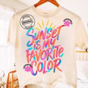 May include: Cream-colored t-shirt with the text "Sunset is my favorite color" in vibrant, colorful lettering. The design includes a sun graphic and floral accents. The shirt is hanging on a wooden hanger.
