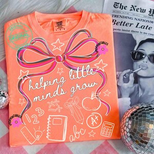 May include: Coral-colored t-shirt with a large bow design in pink, black, and white. The shirt features the phrase "helping little minds grow" and school-themed graphics. A newspaper and disco ball are in the background.