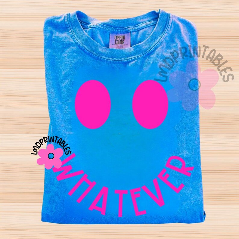 May include: A blue Comfort Colors t-shirt with a smiley face design. The smiley face has two large pink circles for eyes and the word "WHATEVER" in pink letters for the mouth. The shirt is folded and has a tag in the upper left corner.