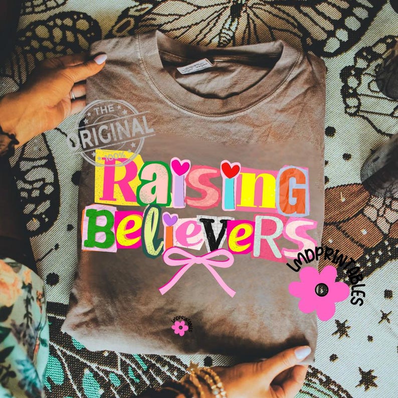 May include: A brown t-shirt with the words "Raising Believers" in colorful, block letters. The design includes hearts, a bow, and the text "The Original". The shirt is folded and held by hands.