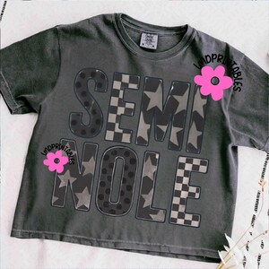 May include: Dark gray t-shirt with the word "SEMINOLE" in a bold, block letter design. The letters feature patterns including polka dots, stars, and a checkered design. A pink flower graphic is on the shirt. The shirt is a Comfort Colors brand.