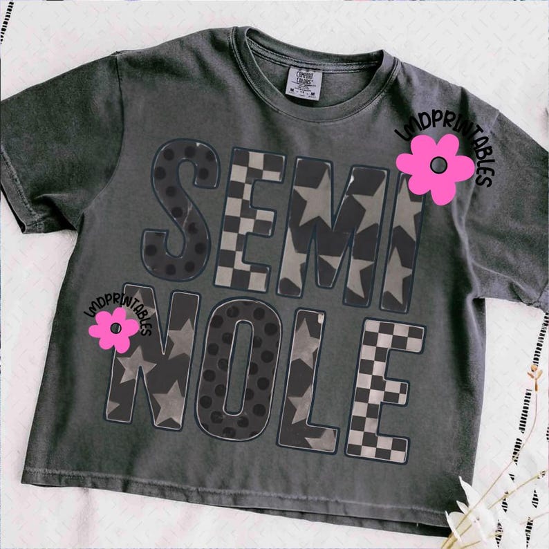 May include: Dark gray t-shirt with the word "SEMINOLE" in a bold, block letter design. The letters feature patterns including polka dots, stars, and a checkered design. A pink flower graphic is on the shirt. The shirt is a Comfort Colors brand.