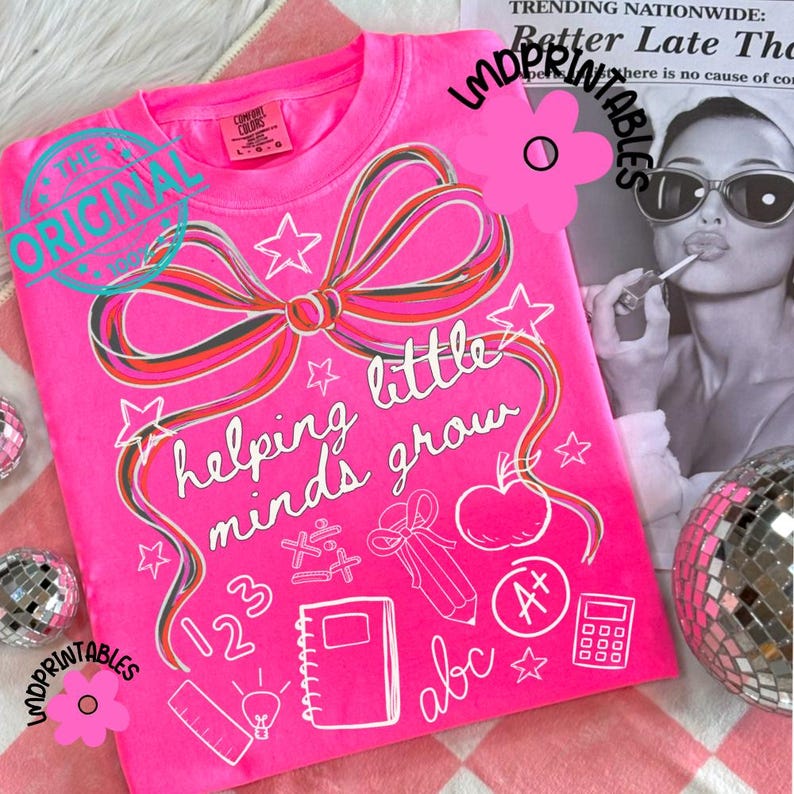 May include: A bright pink t-shirt with a large bow and the words "helping little minds grow" in white. The shirt features school-themed graphics like stars, an apple, and a pencil. The shirt is a Comfort Colors brand.