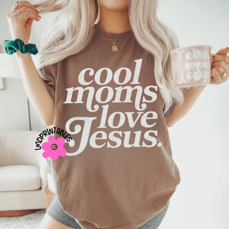Puede incluir: Una camiseta marr&oacute;n con el texto "cool moms love Jesus" en letras blancas. La camiseta es de color neutro y tiene un corte relajado. Una taza rosa con texto se sostiene en la mano derecha.