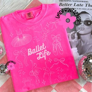 May include: A bright pink sweatshirt with a white graphic design featuring ballet-themed illustrations. The design includes a tutu, ballet shoes, bows, and the words "Ballet Life". The sweatshirt is folded on a pink and white checkered surface.