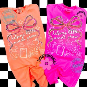 May include: Two t-shirts, one orange and one pink, with the phrase "helping little minds grow" in white script. The design includes a bow, stars, an apple, and school-related icons. The shirts have the word "ORIGINAL" stamped on them.