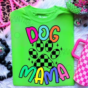 May include: A neon green t-shirt with the words "DOG MAMA" in a colorful, playful font. The design includes a paw print with a checkered pattern. The shirt is a Gildan brand.