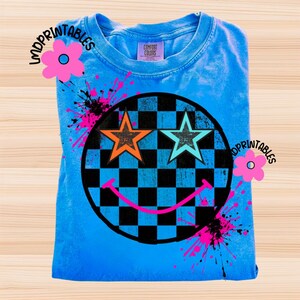 May include: A blue t-shirt featuring a smiley face design. The smiley face has a black and blue chequered background, orange and teal star eyes, and a pink smile. Pink paint splatters and the text "LMDPRINTABLES" are also visible.