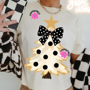May include: Cream-colored t-shirt featuring a Christmas tree design. The tree is gold with black polka dots and a black and white polka dot bow. The text "LMDPRINTABLES" is printed on the shirt with a pink flower.