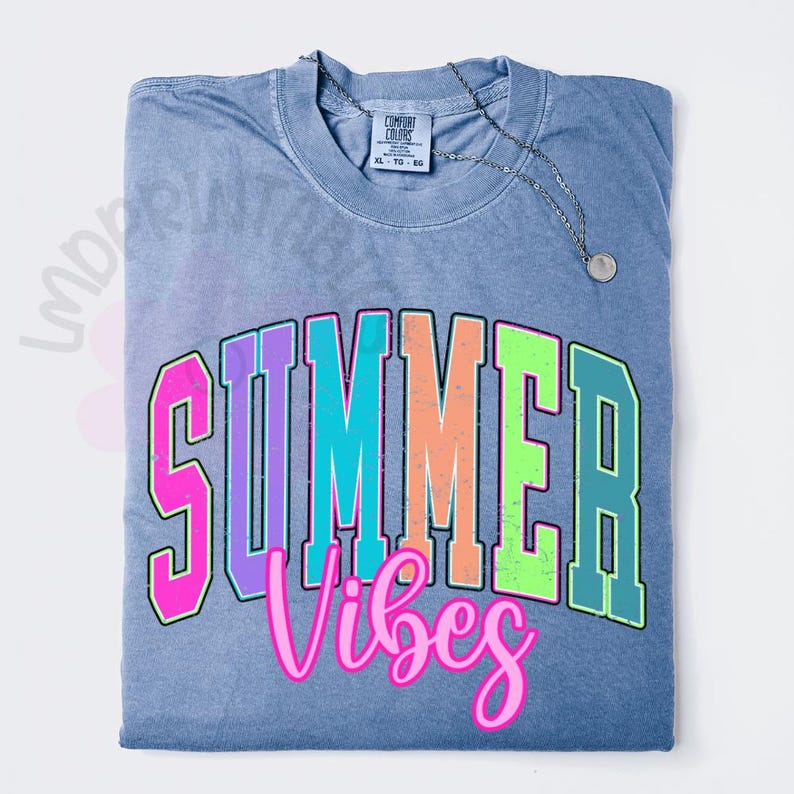 Puede incluir: Una camiseta azul con las palabras "SUMMER Vibes" en un dise&ntilde;o arqueado y colorido. El texto est&aacute; delineado en negro y presenta colores rosa, morado, naranja, verde y turquesa. Un collar plateado con un colgante descansa sobre la camiseta.