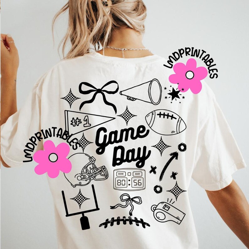 May include: Cream-colored t-shirt with a black graphic design featuring football-themed elements. The design includes a football, helmet, megaphone, and the words "Game Day." Pink flower and text "LMDPRINTABLES" are also visible.