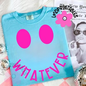 May include: A light blue t-shirt with a smiley face design. The smiley face features two large pink circles for eyes and the word "WHATEVER" in pink below. The shirt is a Comfort Colors brand.