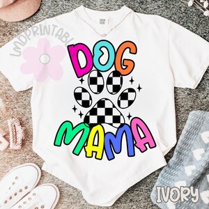 May include: White t-shirt with a colorful graphic that says "DOG MAMA". The design includes a paw print with a checkered pattern and the word "DOG" in pink, teal, and orange. The word "MAMA" is in green, blue, and pink.