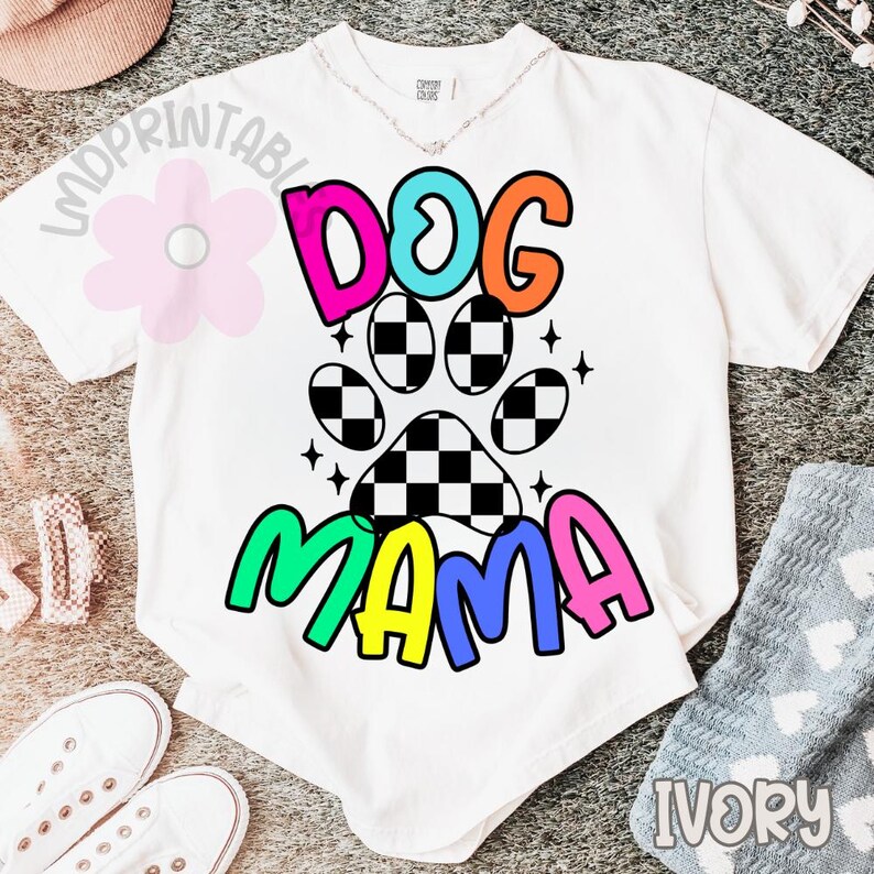May include: White t-shirt with a colorful graphic that says "DOG MAMA". The design includes a paw print with a checkered pattern and the word "DOG" in pink, teal, and orange. The word "MAMA" is in green, blue, and pink.