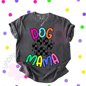 May include: Dark gray t-shirt with the words "DOG MAMA" in bright, colorful letters. The design includes a paw print with a checkered pattern. A silver necklace with a small pendant is visible.