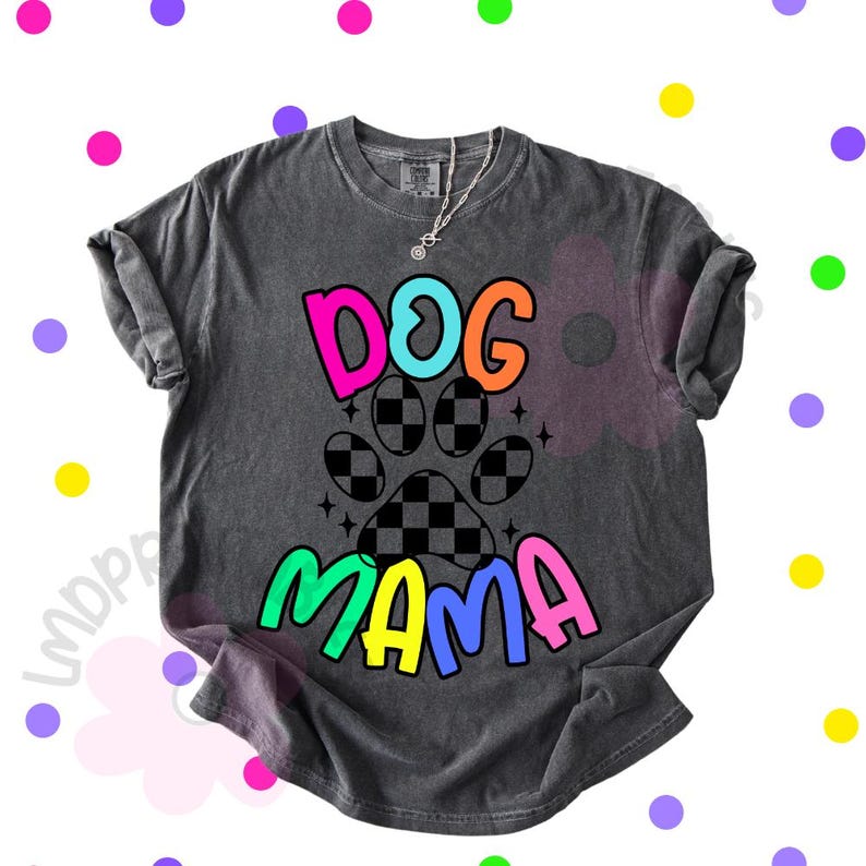 May include: Dark gray t-shirt with the words "DOG MAMA" in bright, colorful letters. The design includes a paw print with a checkered pattern. A silver necklace with a small pendant is visible.