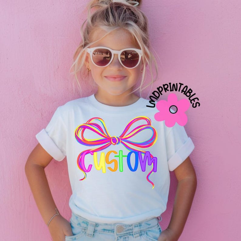 May include: A white t-shirt featuring a colorful bow design with the word "CUSTOM" in rainbow colors. The shirt is worn by a young person wearing sunglasses, against a pink background. The design is playful and vibrant.