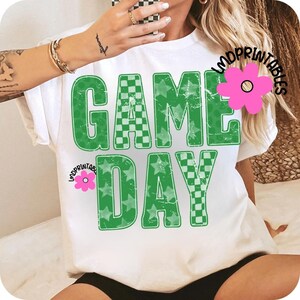 May include: White t-shirt with the words "GAME DAY" in large, distressed green letters. The letters are filled with a checkerboard pattern and stars. The shirt has a relaxed fit and short sleeves.