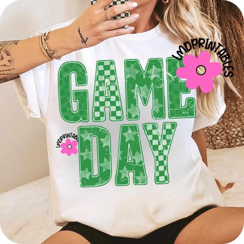 May include: White t-shirt with the words "GAME DAY" in large, distressed green letters. The letters are filled with a checkerboard pattern and stars. The shirt has a relaxed fit and short sleeves.