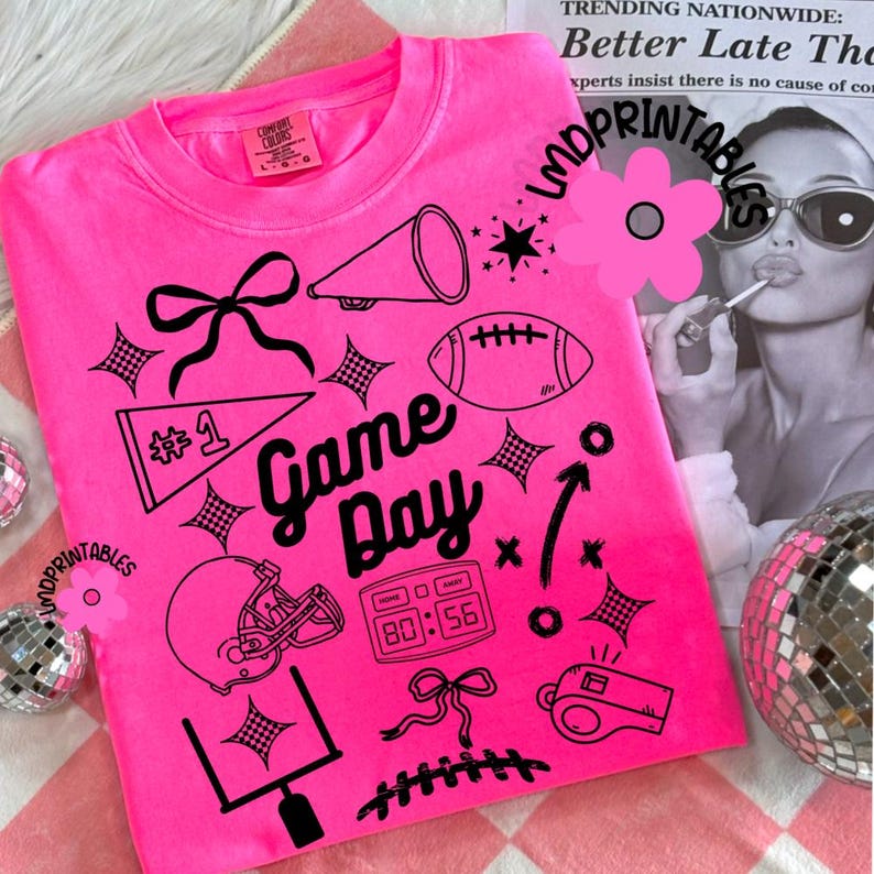 May include: Bright pink sweatshirt with black graphic elements. The design includes a football, helmet, megaphone, and the words "Game Day." Additional details include a scoreboard, bow, and stars. The shirt has a crew neck and long sleeves.