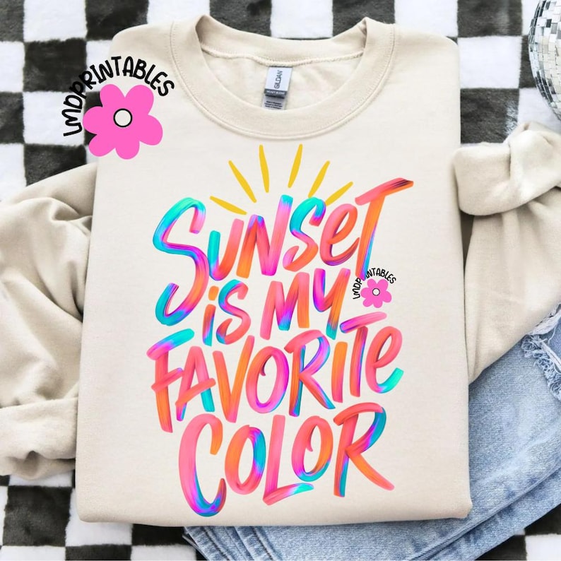 May include: Cream-colored sweatshirt with the text "Sunset is my favorite color" in vibrant, colorful lettering. The design features a sunburst and a pink flower graphic. The sweatshirt is a casual, comfortable style.