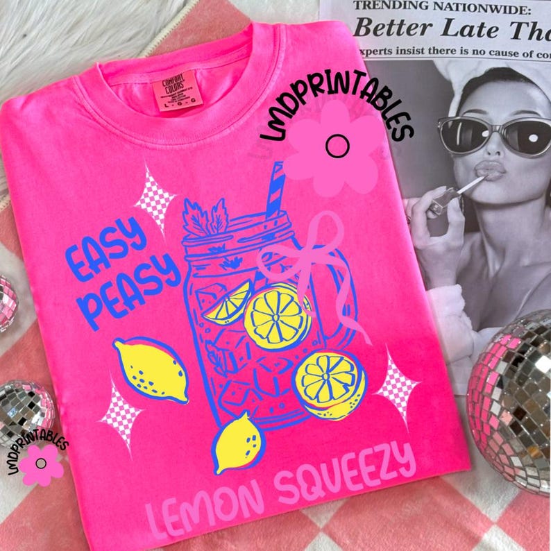 May include: A vibrant pink sweatshirt featuring a lemonade design with lemons and the text "EASY PEASY LEMON SQUEEZY" in blue and yellow. The long-sleeved top has a crew neck. The brand name "LMDPRINTABLES" is also visible.